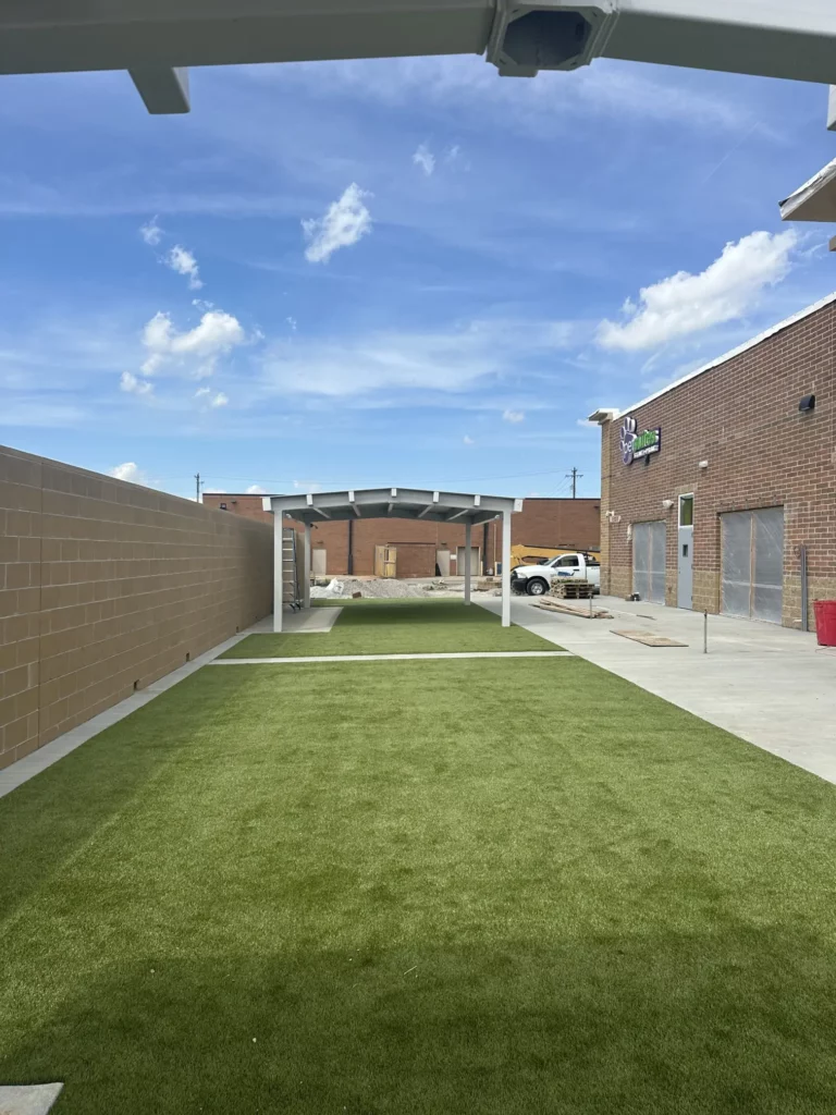 K9Grass artificial turf play yard with covered pergola structure at PetSuites Indianapolis under construction, brick exterior visible.