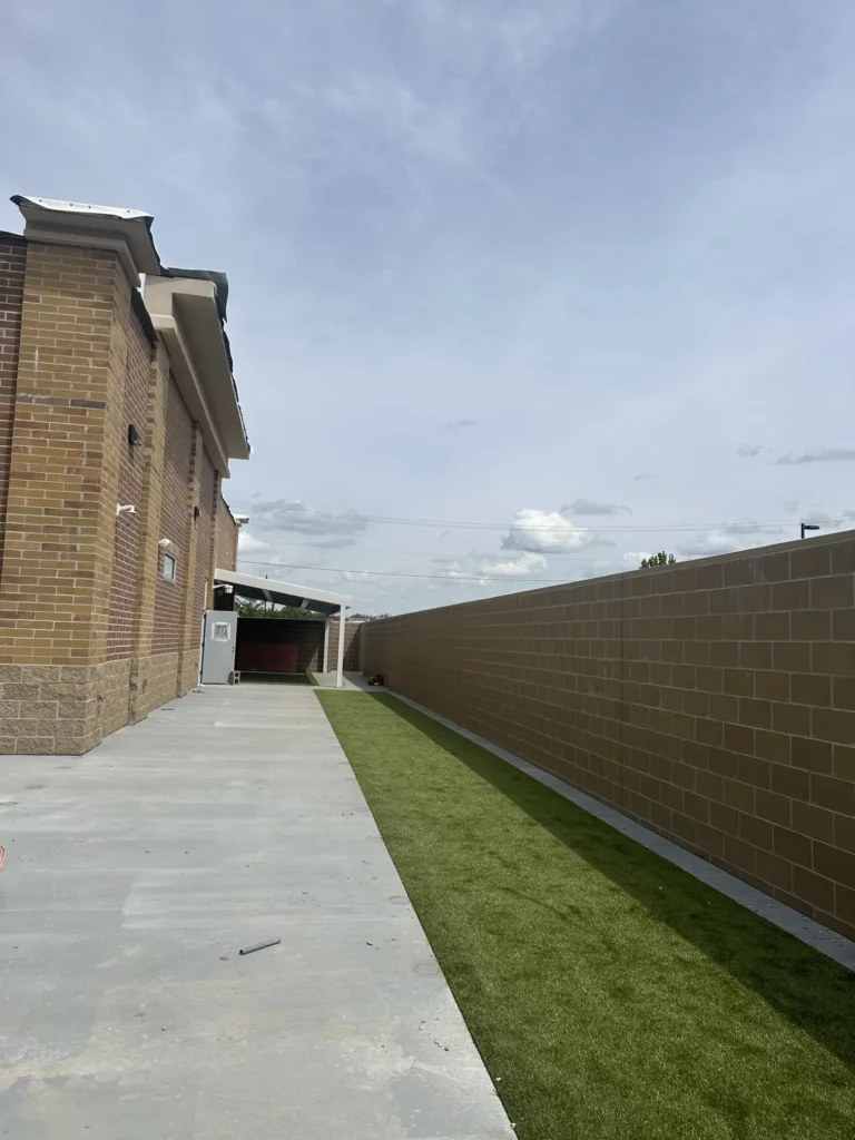 Artificial K9Grass turf strip alongside concrete walkway between brick building and block retaining wall at PetSuites Indianapolis.