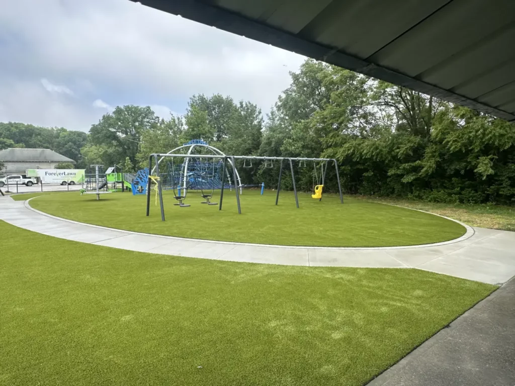 Lush artificial turf playground at Municipal Gardens Indianapolis with swing sets, climbing structure, and curved concrete border.