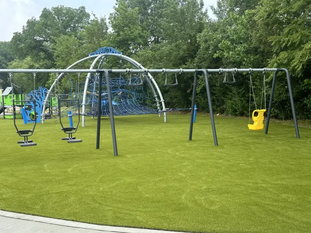 Artificial turf playground at Municipal Gardens Indianapolis with swing sets, rope climbing dome, and colorful play equipment.