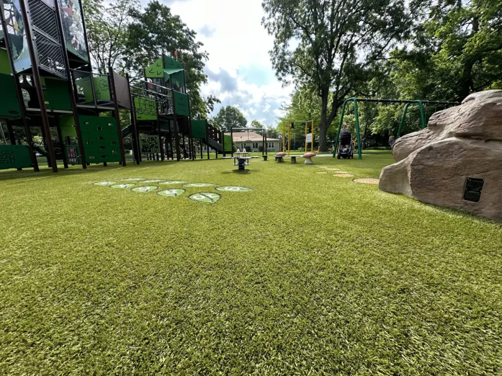 Modern municipal playground with Playground Grass Ultra artificial turf, large climbing structure, swings, and decorative boulder in a tree-lined park.