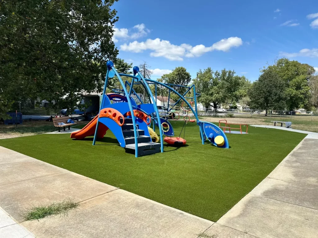 Colorful blue playground equipment on lush artificial turf at Indianapolis municipal park, surrounded by trees under blue sky.