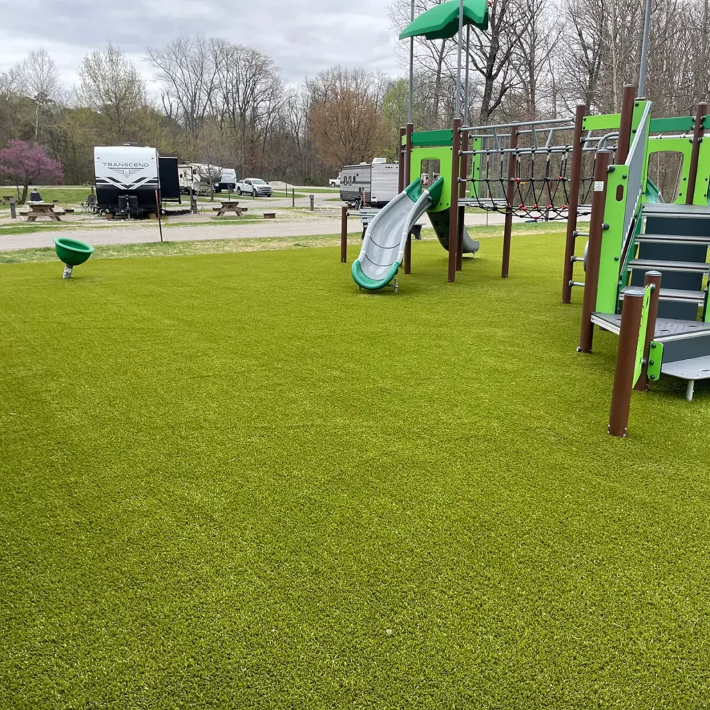 Lush artificial turf surrounds a green and brown playground structure with slide at an RV campground in early spring.