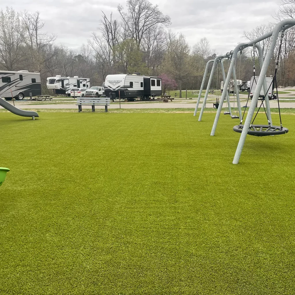 Lush artificial turf playground at RV campground with swing set, slide, and bench surrounded by parked travel trailers.