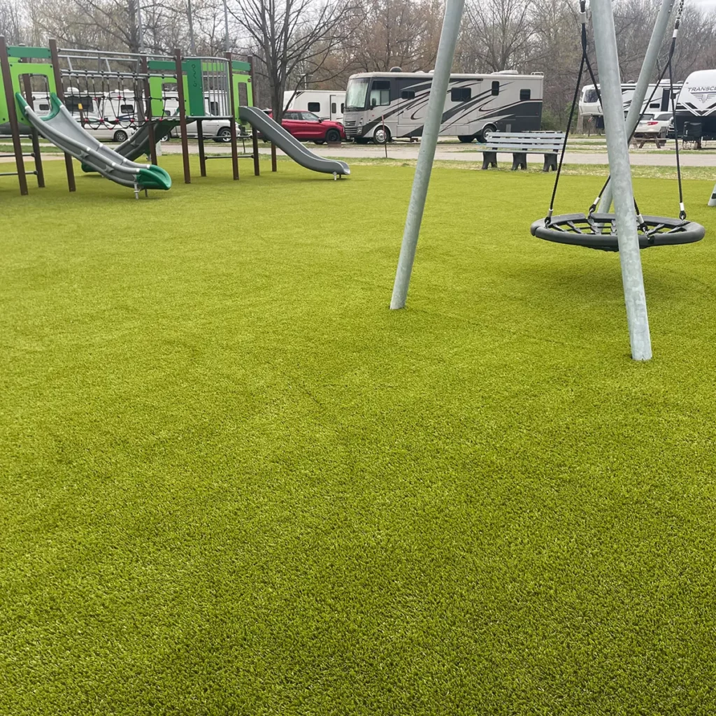 Artificial turf playground at RV campground with swing set, slides, and climbing structure on lush green synthetic grass.