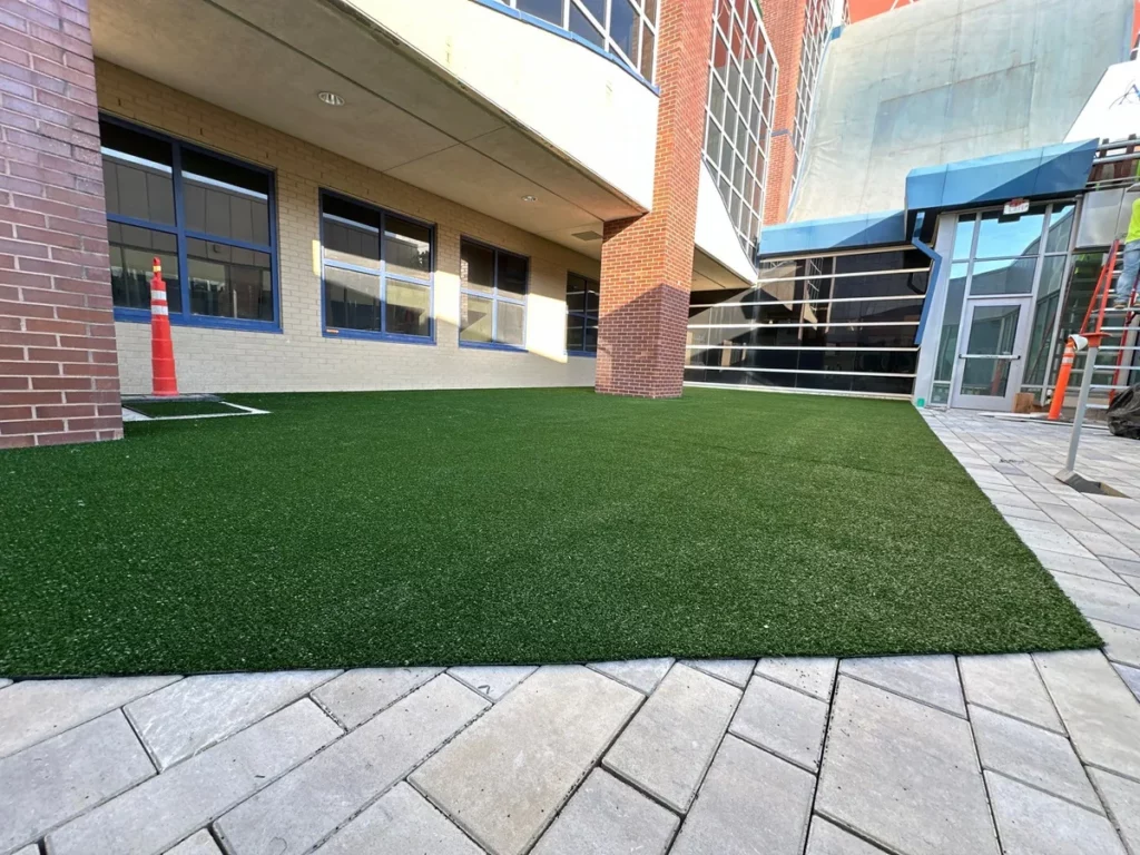 Playground Grass Explore artificial turf installed in hospital courtyard beside brick building with stone paver border, construction ongoing.