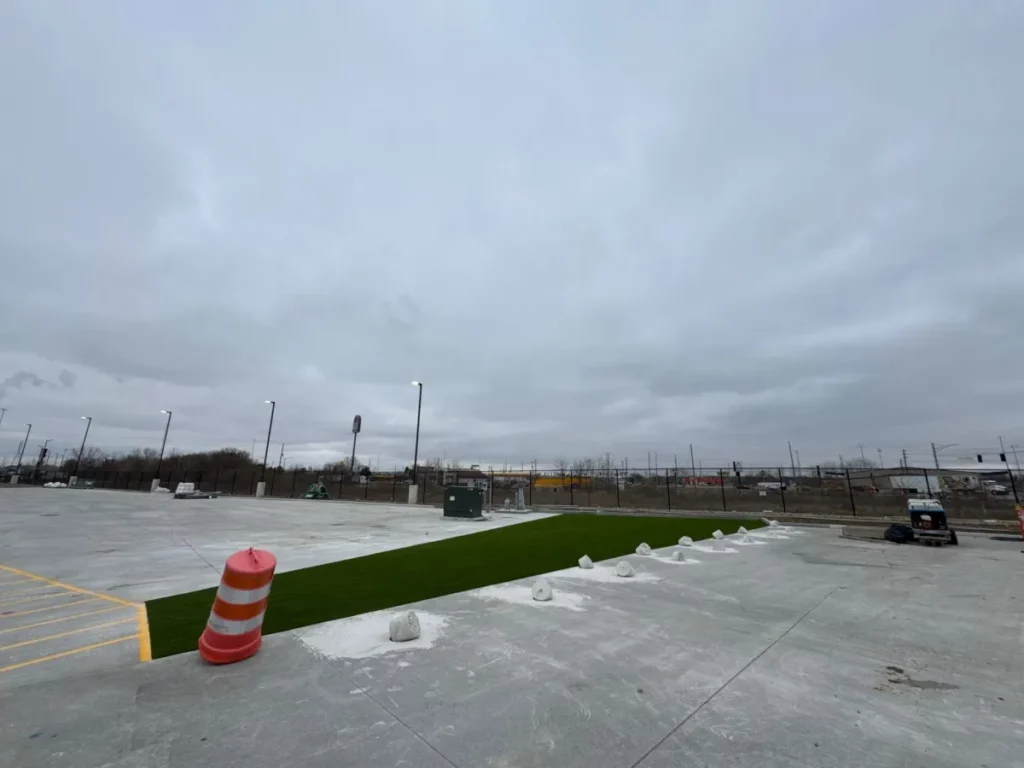 Newly installed K9Grass Classic+ artificial turf strip in concrete dog play area with boulders, fencing, and light poles under overcast sky.