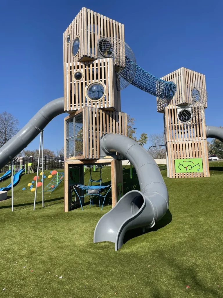 Playground Grass Ultra artificial turf beneath towering wooden tower playground with tube slides and net tunnels at municipal park.