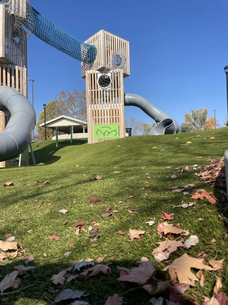 Playground Grass Ultra artificial turf on hillside playground with tube slides and wooden tower structure, fall leaves, clear sky.