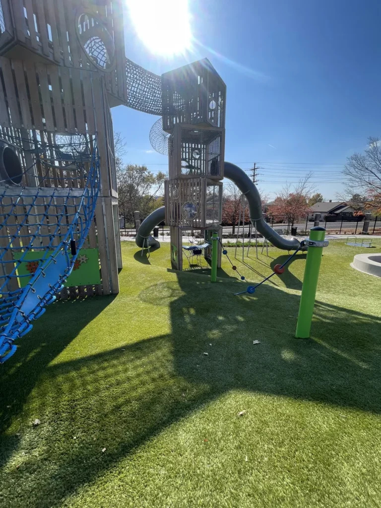 Playground Grass Ultra artificial turf installed at municipal park with multi-level wood tower, tube slides, rope climbers, and blue net structures.