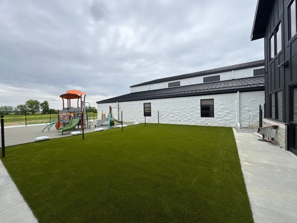 Playground Grass Ultra artificial turf installed beside modern school building with colorful playground equipment on rubber mulch surface.