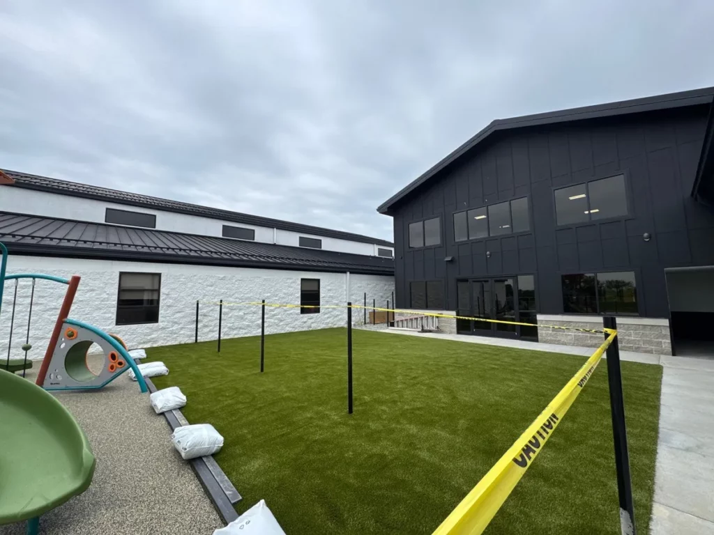 Freshly installed Playground Grass Ultra artificial turf in school courtyard with playground equipment, caution tape marking new construction zone.