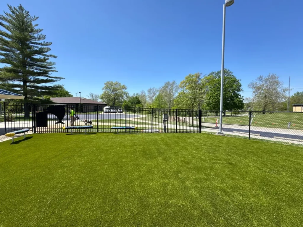 Bright green Playground Grass Ultra artificial turf installed at fenced municipal park playground with light pole and building backdrop.