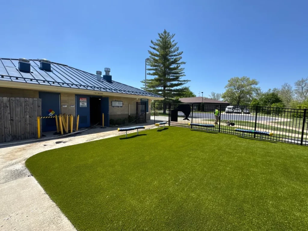 Bright green Playground Grass Ultra artificial turf installed around a municipal pool facility with black metal fencing and benches.