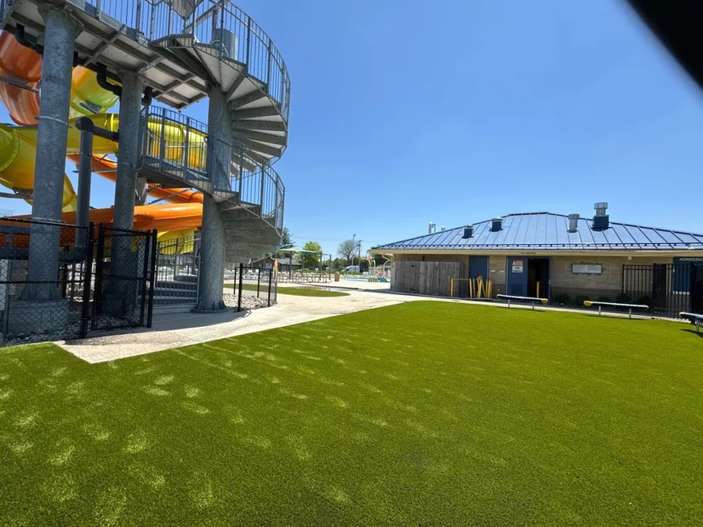 Bright green Playground Grass Ultra artificial turf installed beside a water park slide tower and brick admission building on a sunny day.