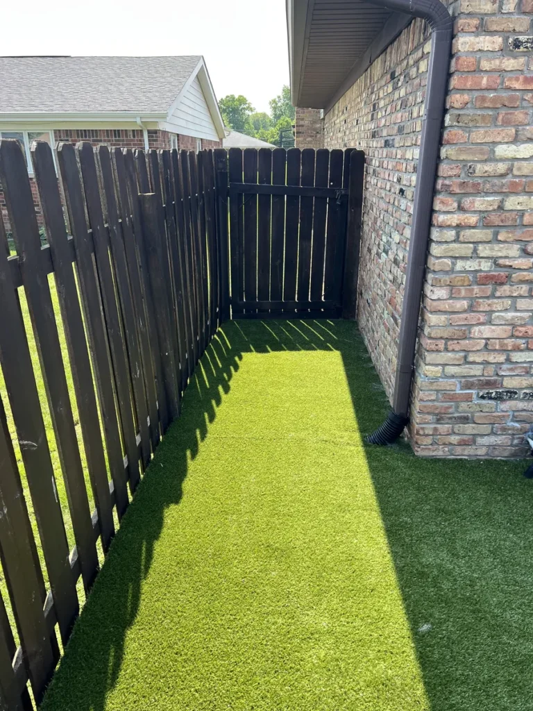 K9Grass Elite artificial turf installed in narrow side yard between brick home and dark wood fence with gate.