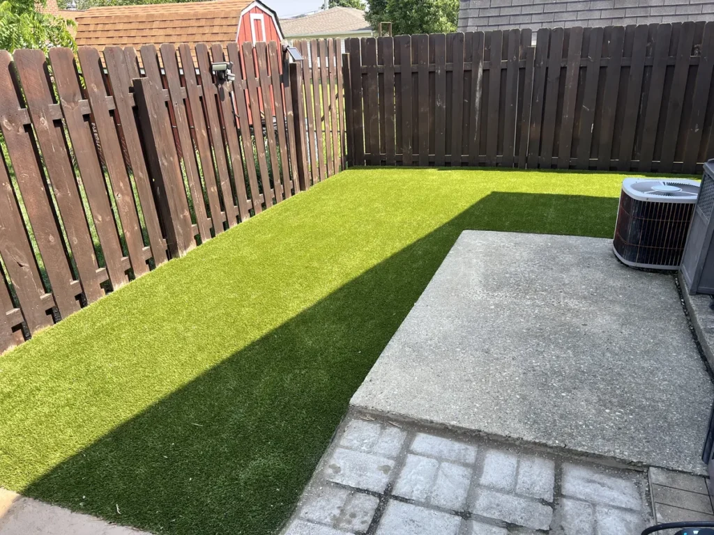 K9Grass Elite artificial turf installed in fenced backyard pet area alongside concrete patio with AC unit nearby.