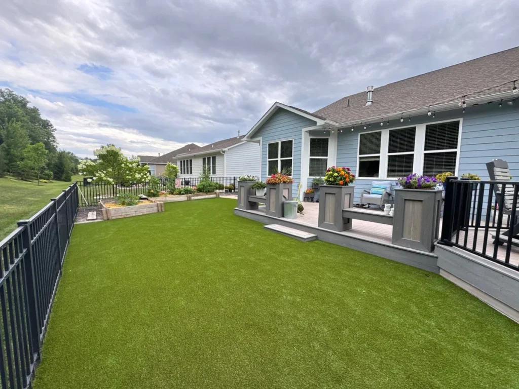 K9Grass Endura artificial turf installed in fenced suburban backyard beside gray deck with flower planters and raised garden beds.