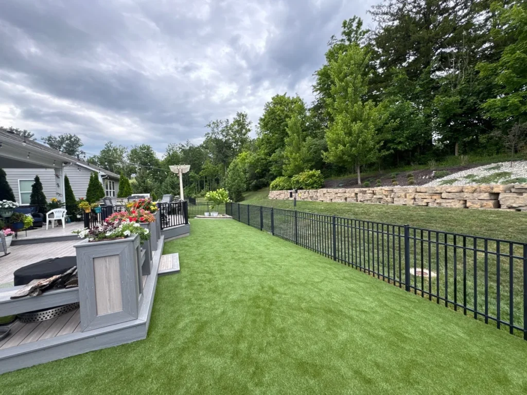K9Grass Endura artificial turf pet yard with black aluminum fence, stone retaining wall, and gray deck with flower planters.