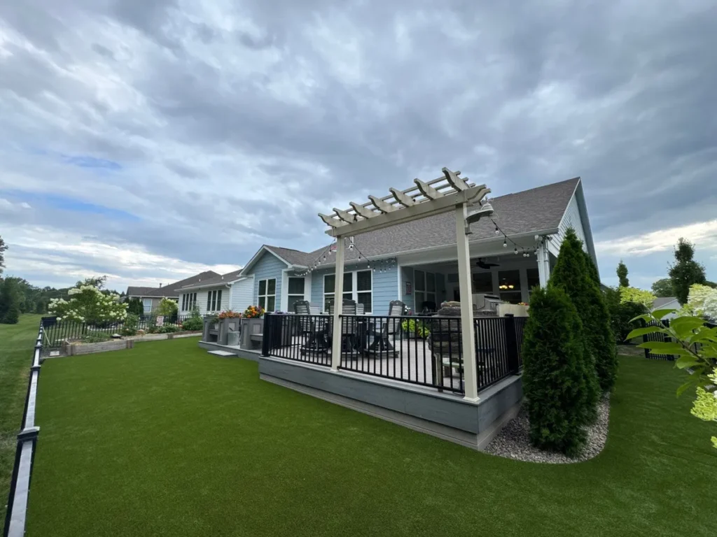 K9Grass Endura artificial turf installed in fenced backyard beside gray composite deck with pergola and string lights.