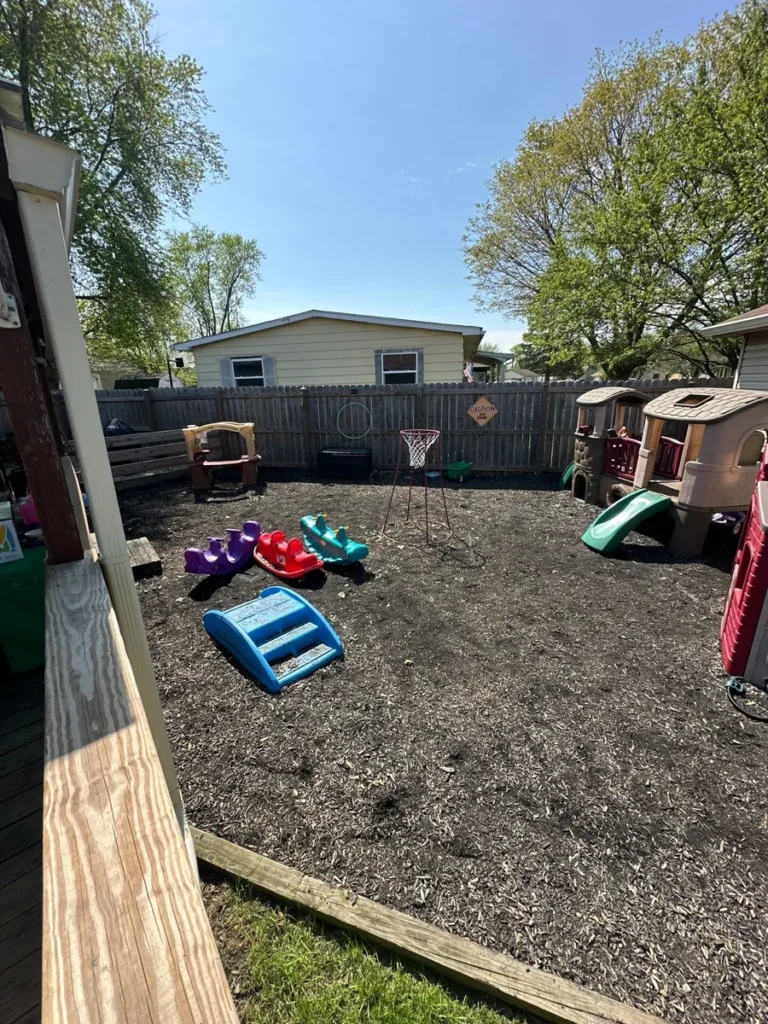 Backyard playground with wood chip mulch ground cover, colorful toddler toys, basketball hoop, and plastic playhouse structure.