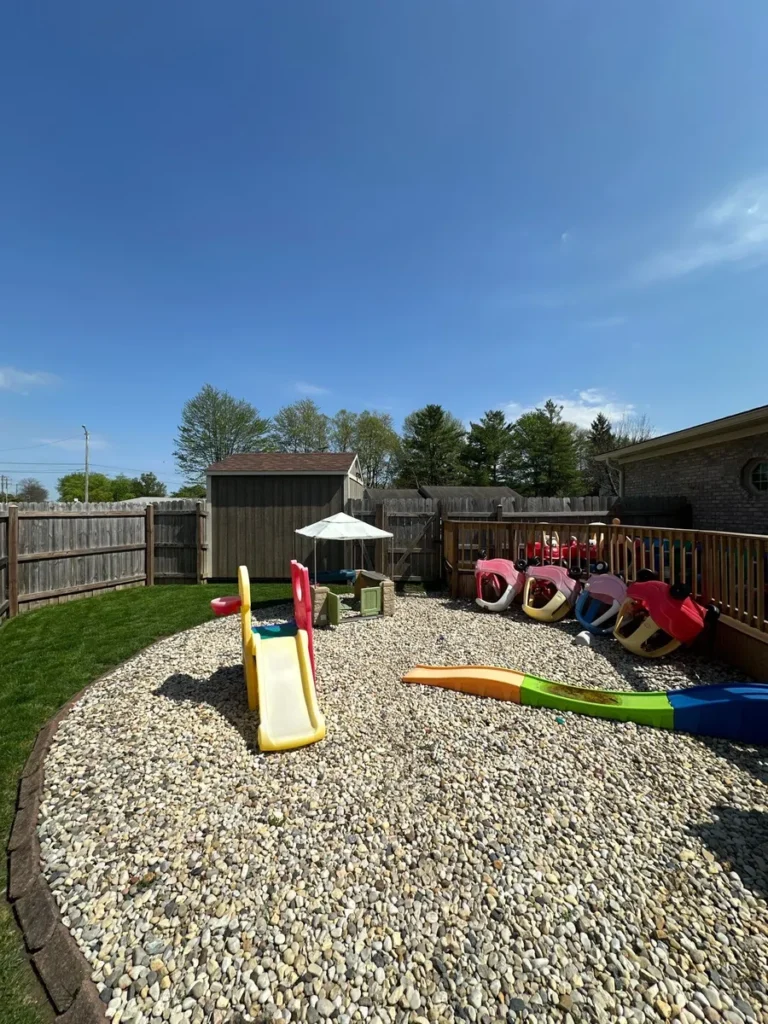 Backyard playground with pea gravel safety surface, colorful toddler slides, ride-on toys, wood deck, and storage shed.