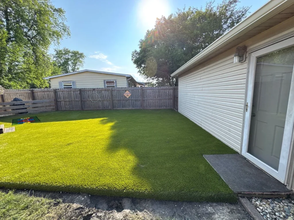 Playground Grass Ultra artificial turf installed in residential backyard near school, vibrant green surface, wood fence, sunny day.