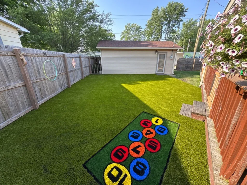 Backyard school playground with Playground Grass Ultra artificial turf, colorful numbered hopscotch mat, wood fence, and detached garage.