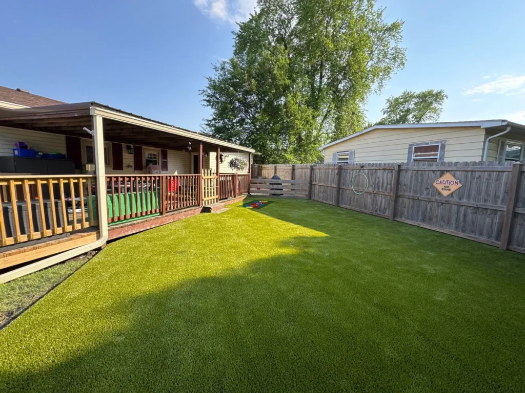 Lush artificial playground turf installed in fenced residential backyard with wood deck, "Caution Kid Zone" sign, and cornhole game.