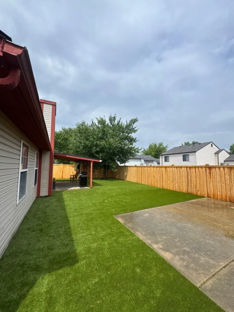 Lush artificial K9Grass pet turf installed in fenced suburban backyard with concrete patio, covered patio area, and mature shade tree.