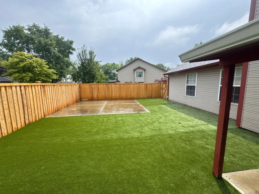 Fenced suburban backyard with K9Grass artificial turf installation beside home, concrete patio pad, wood privacy fence.