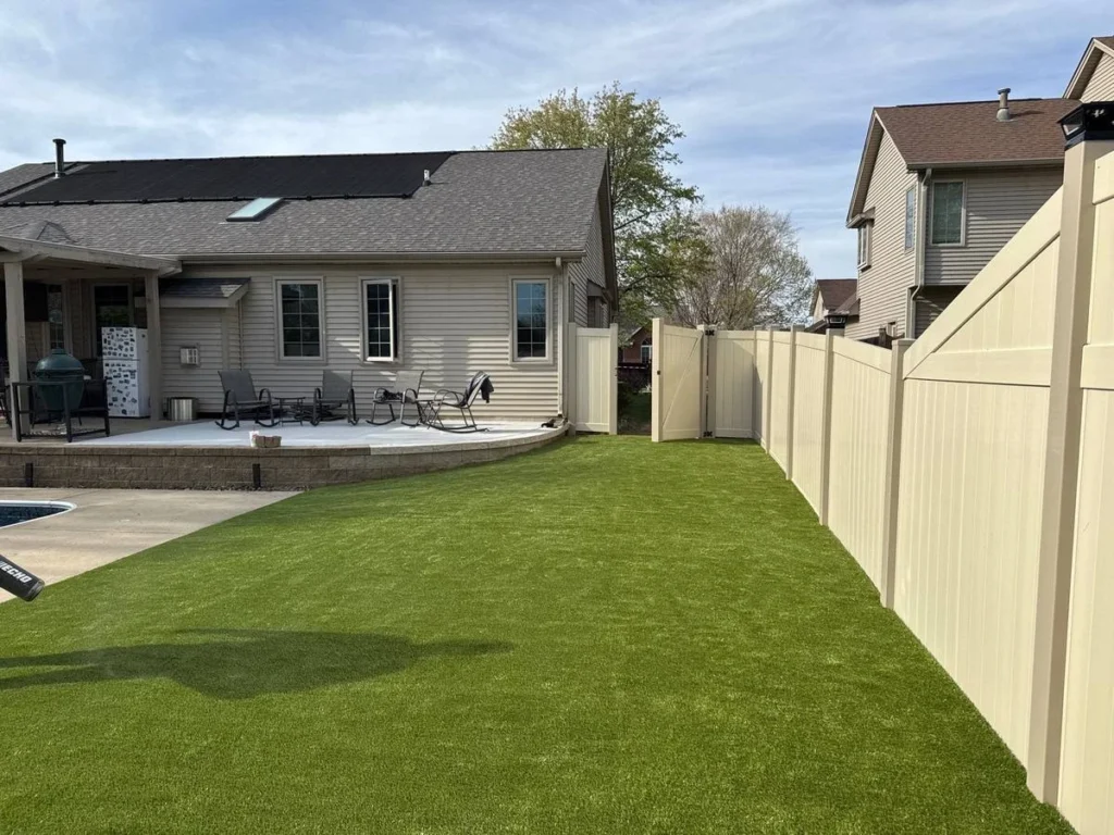 Lush artificial K9Grass pet turf installed in fenced suburban backyard beside patio, pool, and covered outdoor living area.