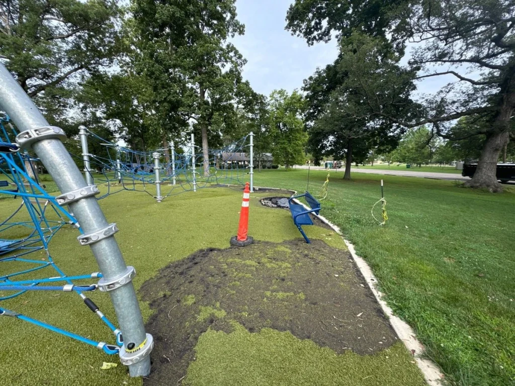 Burned artificial turf repair area with orange safety cone near blue rope climbing structure in municipal park playground.