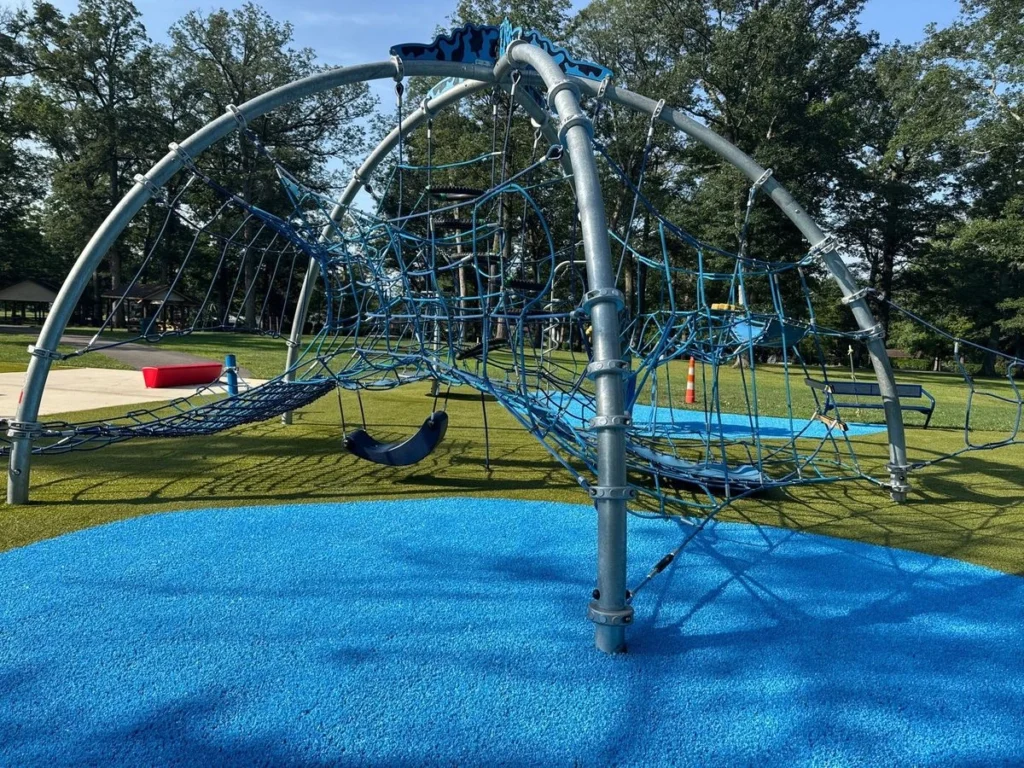 Blue synthetic turf surrounds a large rope climbing dome structure at a municipal park playground in New Castle.