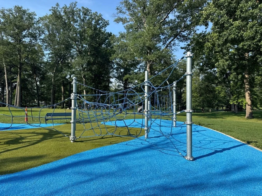 Blue rubber safety surfacing surrounds a rope climbing structure in a lush municipal park with mature trees.