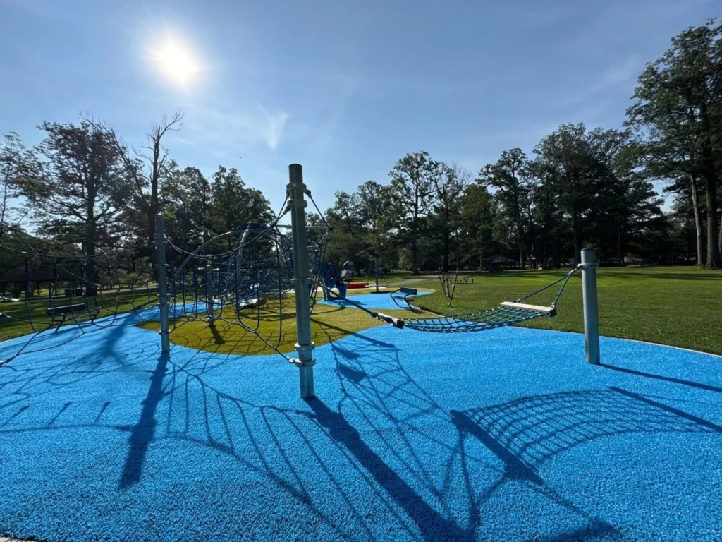 Blue rubber safety surfacing surrounds rope climbing structures at a restored municipal playground in New Castle, sunny park setting.
