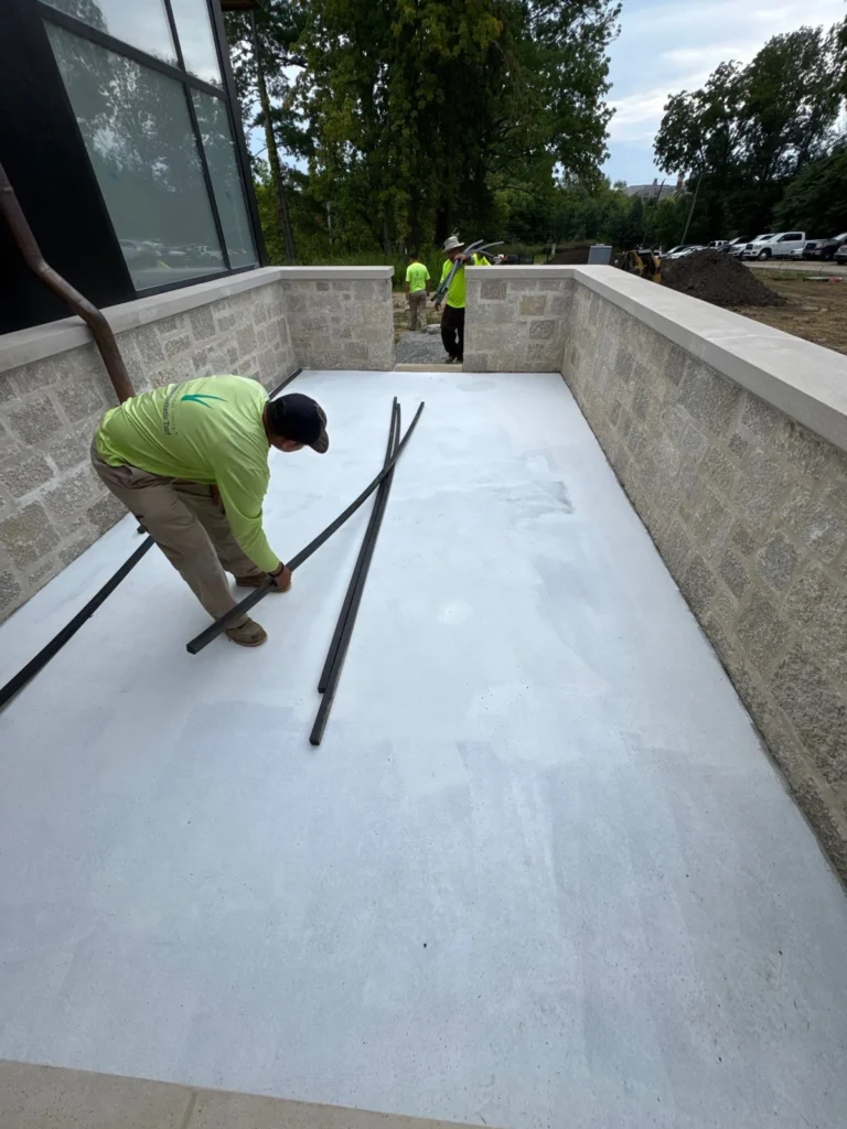 Workers in lime green shirts install K9Grass Classic+ pet turf in a stone-walled enclosed yard beside a modern building.