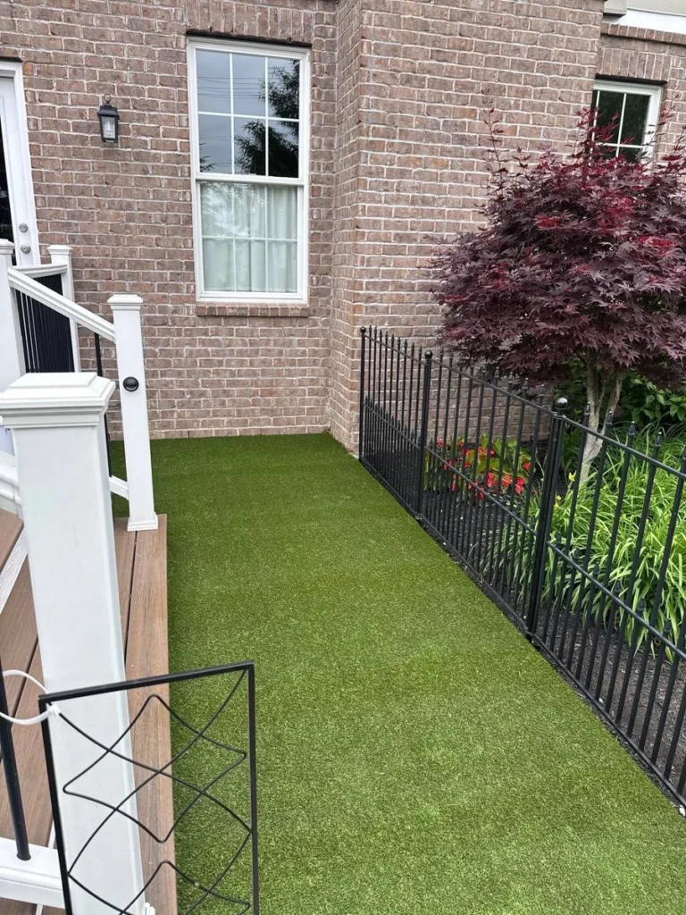 K9Grass artificial pet turf installed in narrow side yard beside brick home with black iron fence and white deck railing.