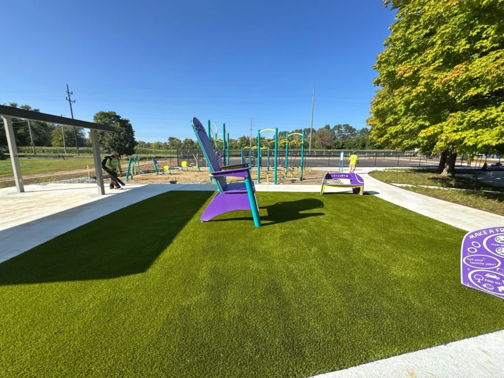 Bright green artificial turf surrounds colorful purple Adirondack chair at newly installed Franklin Township Park playground under blue sky.