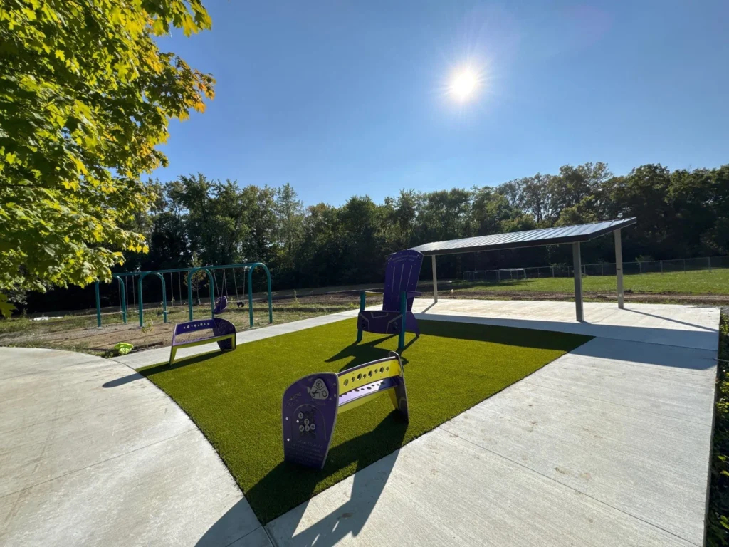 Playground Grass Ultra artificial turf installed under purple benches and chair at Franklin Township Park with swing set and pavilion beyond.