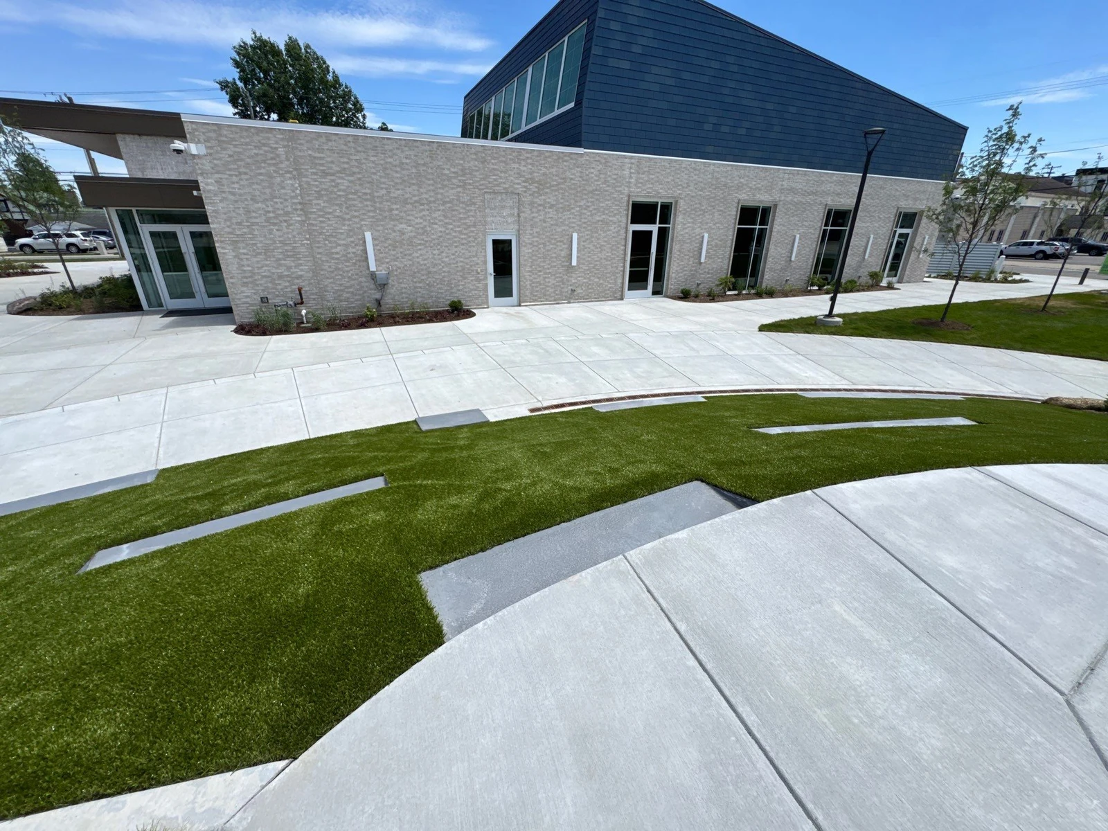 Modern municipal plaza with synthetic Playground Grass turf panels between concrete pavers outside a brick and steel event building.