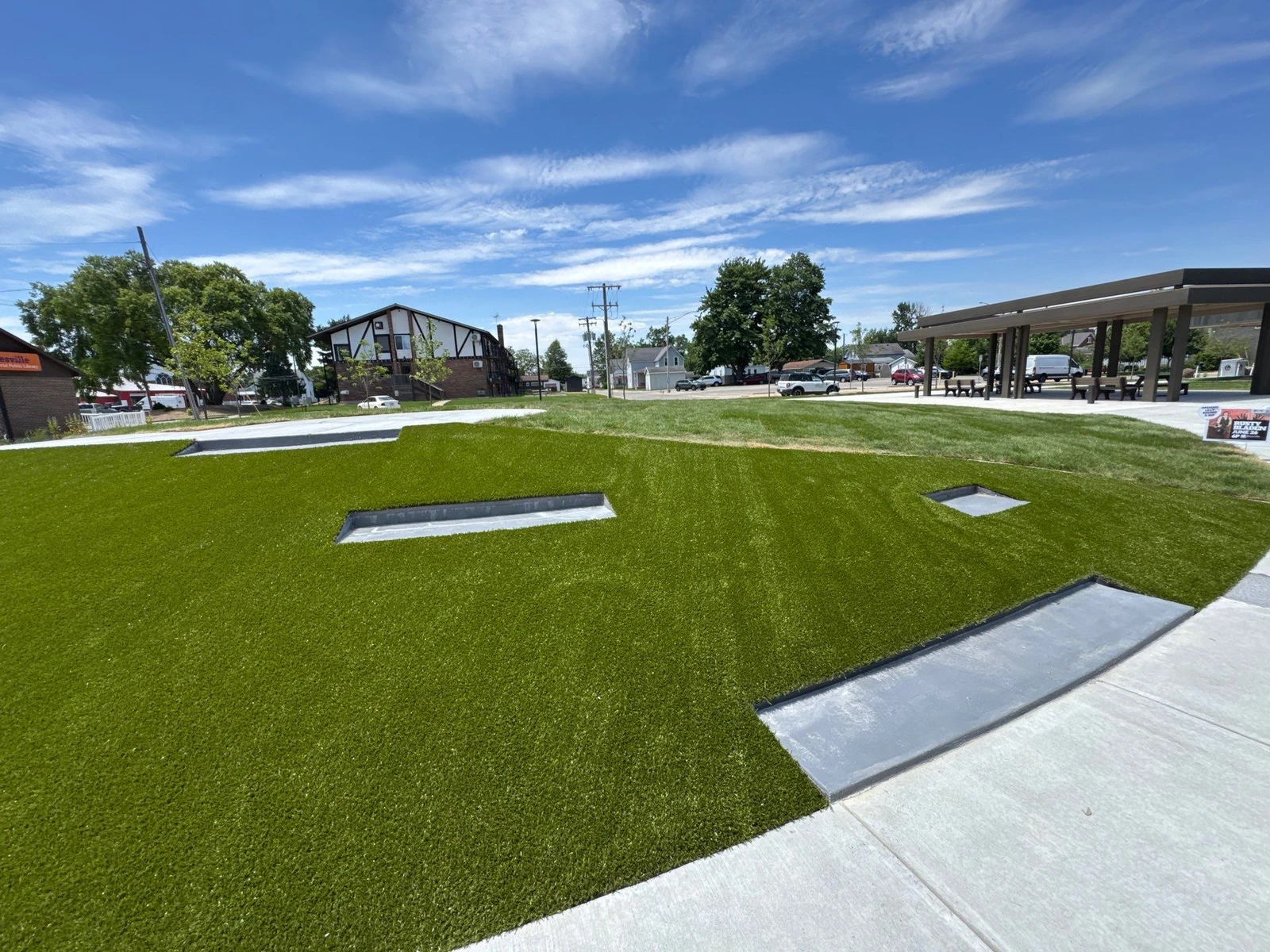 Vibrant artificial playground turf installed at Batesville municipal park with concrete edging, open pavilion, and residential backdrop.