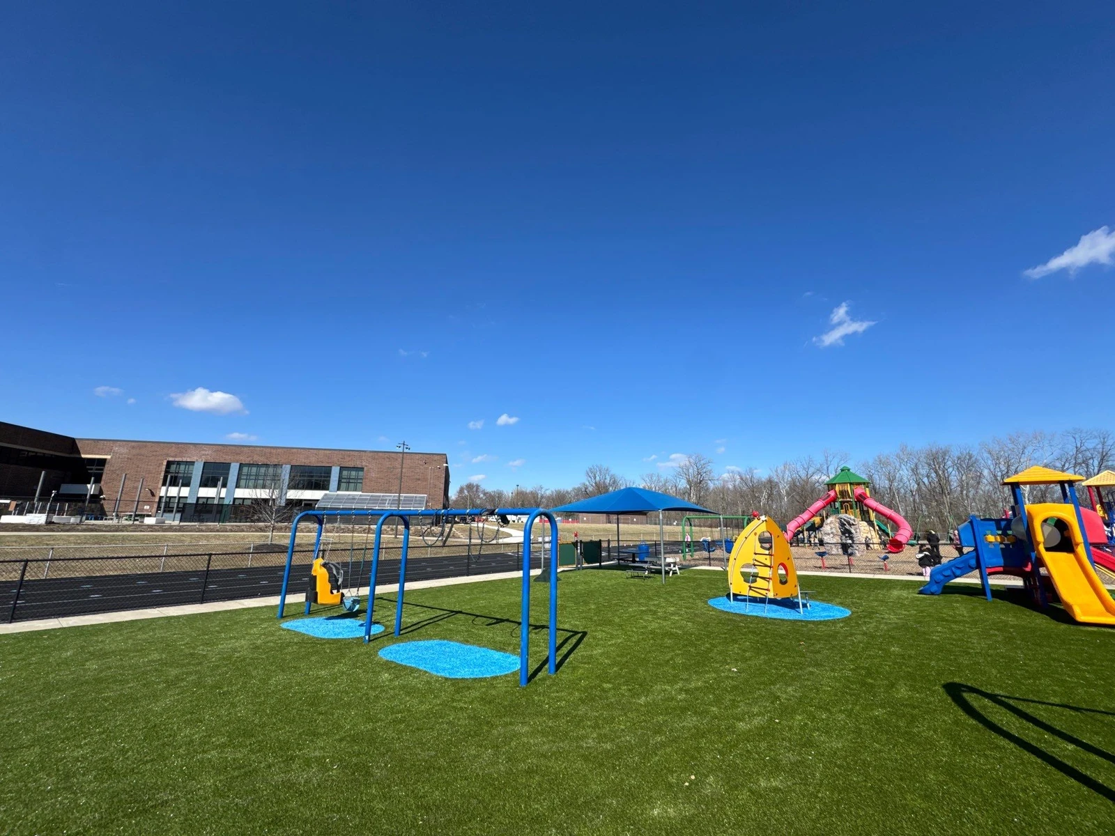 Colorful school playground with artificial turf, blue swing set, slides, and shade canopy beside a running track in Plainfield, IN.