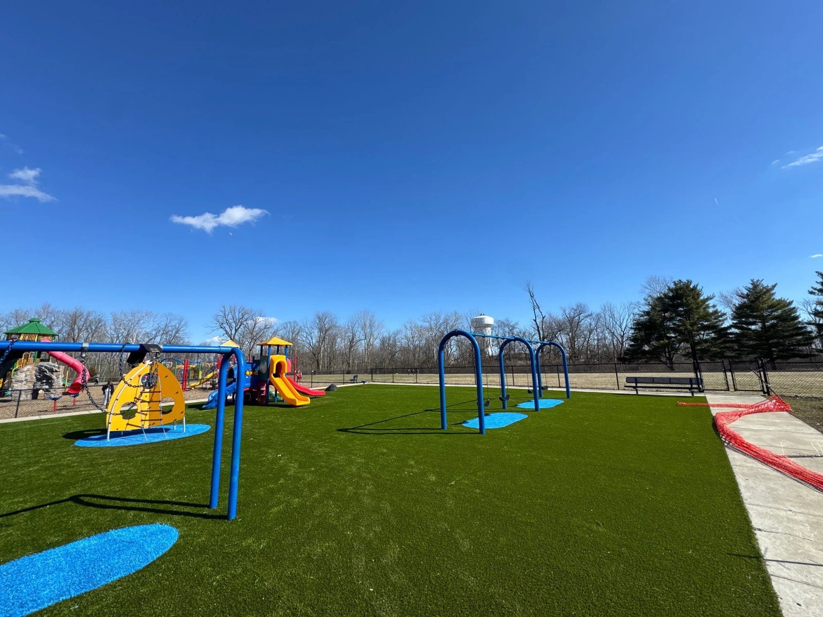 Colorful municipal playground on lush artificial turf with blue arch climbers, slides, and rubber safety pads under clear blue sky.