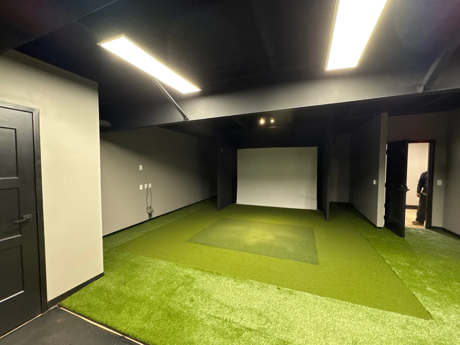 Indoor golf simulator room with GolfGreens artificial turf flooring, dark walls, projection screen, and recessed lighting.