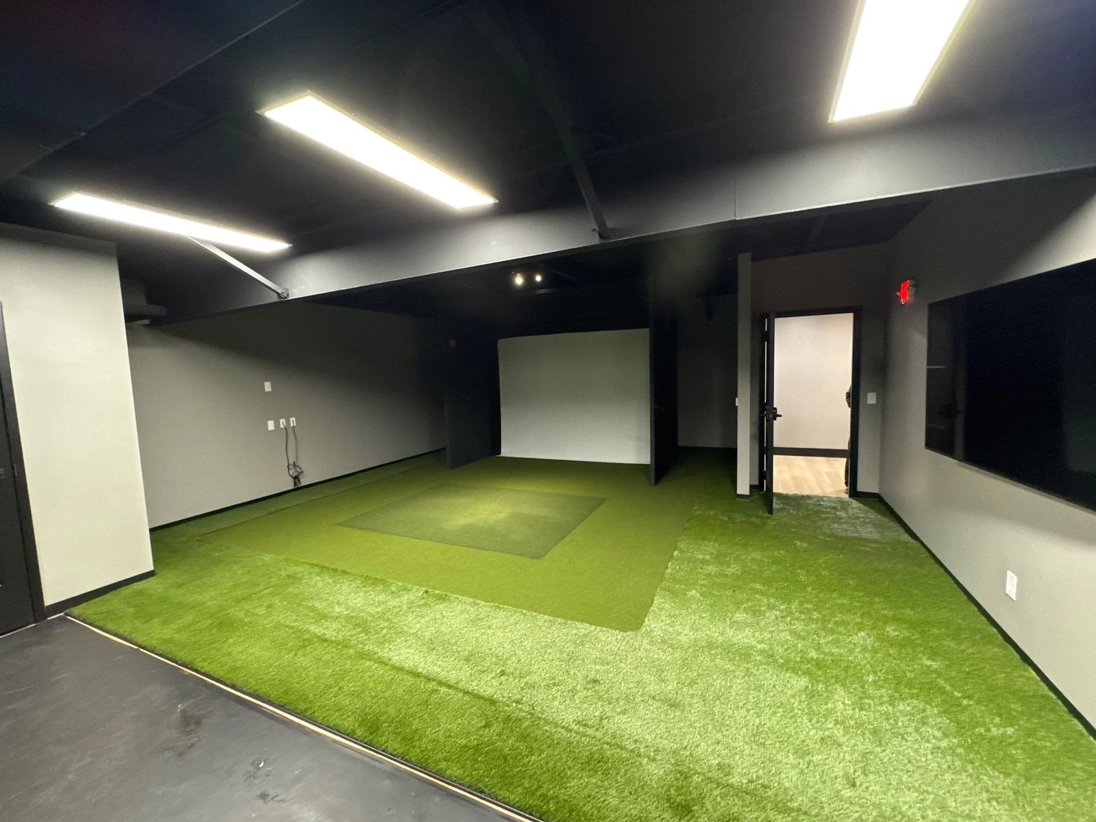 Indoor golf simulator bay with GolfGreens artificial turf flooring, dark walls, overhead lighting, and projection screen.
