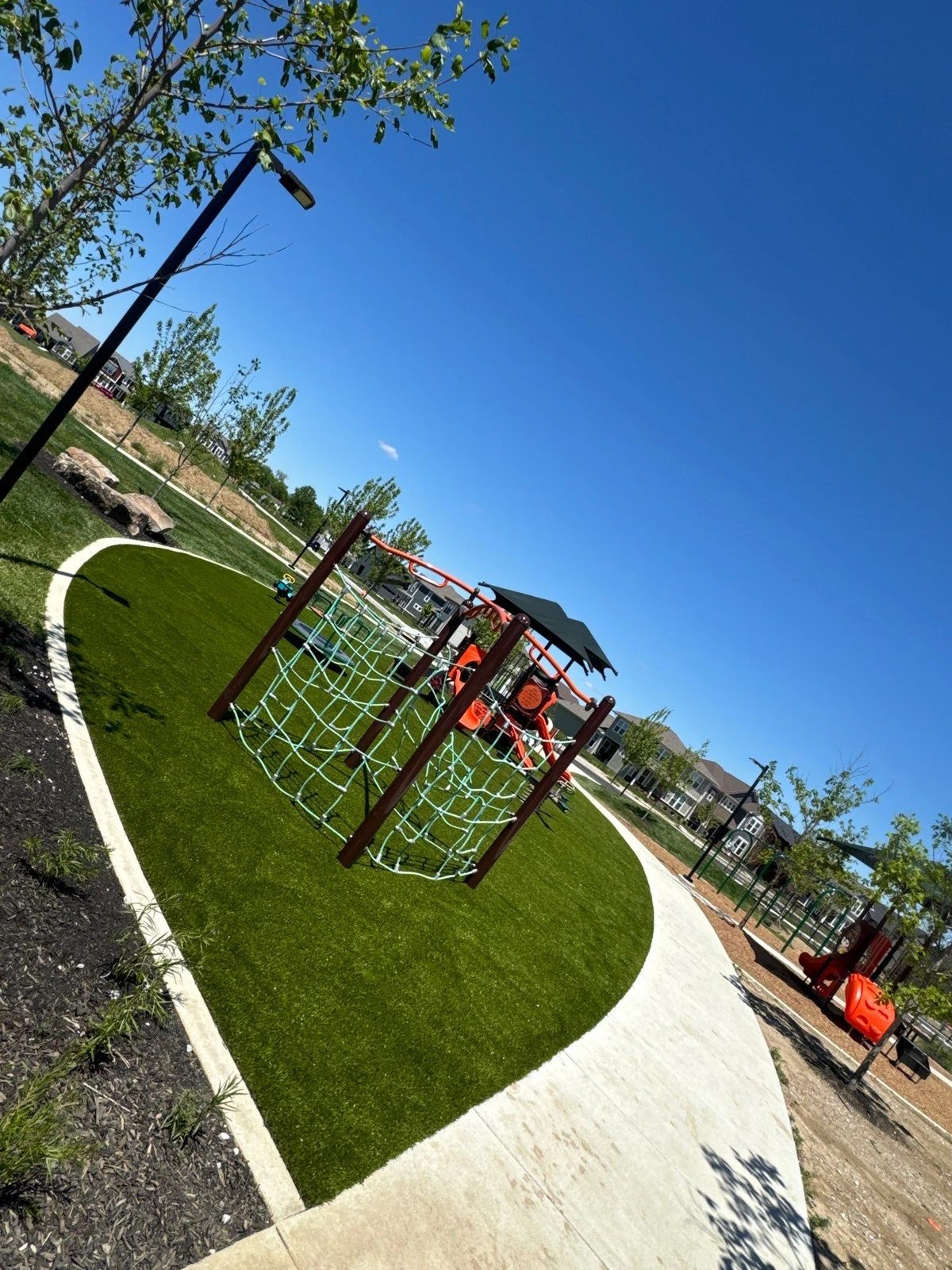 ForeverLawn Playground Grass under colorful climbing structures and play equipment at Hobbs Park, Plainfield IN, bordered by concrete path.