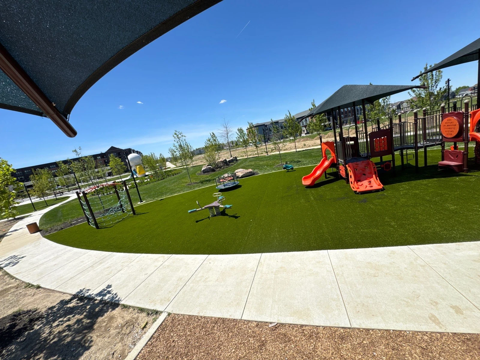 Modern municipal playground with orange play structures, shade sails, and lush artificial Playground Grass turf in Plainfield, IN.