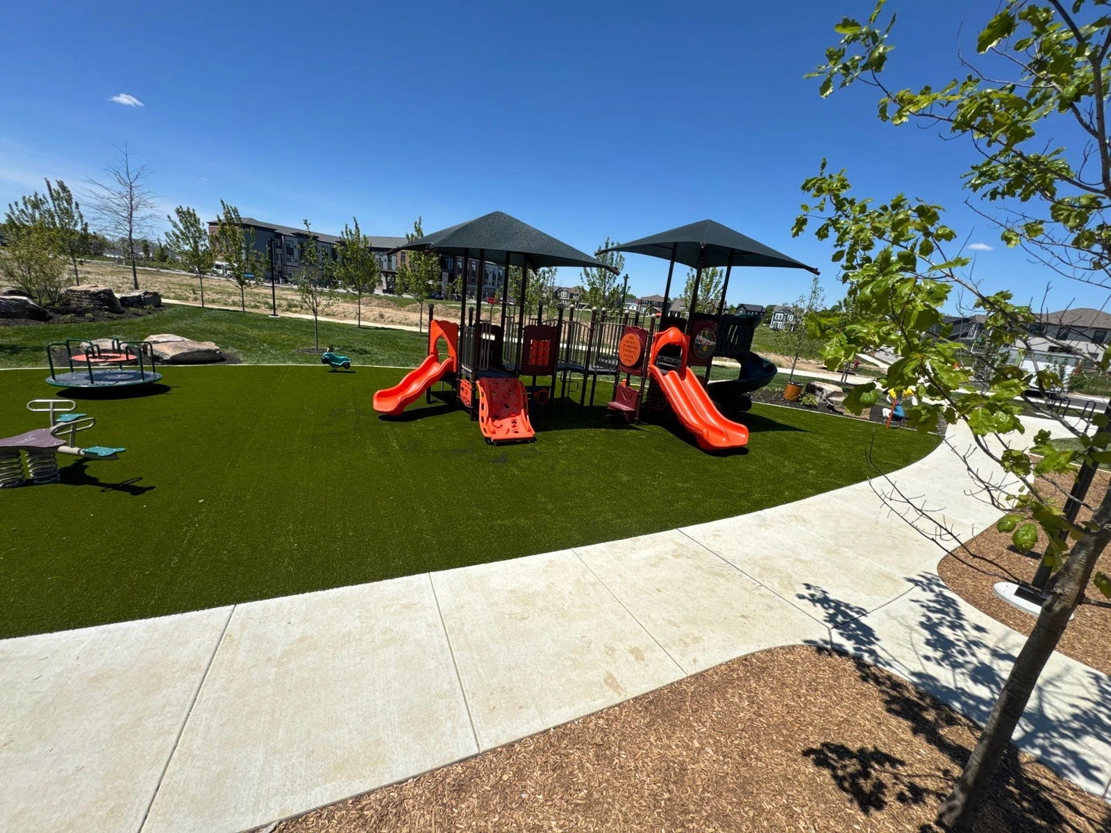 Modern municipal playground with orange slides on lush artificial turf, shaded play structure, and concrete path, Plainfield IN.