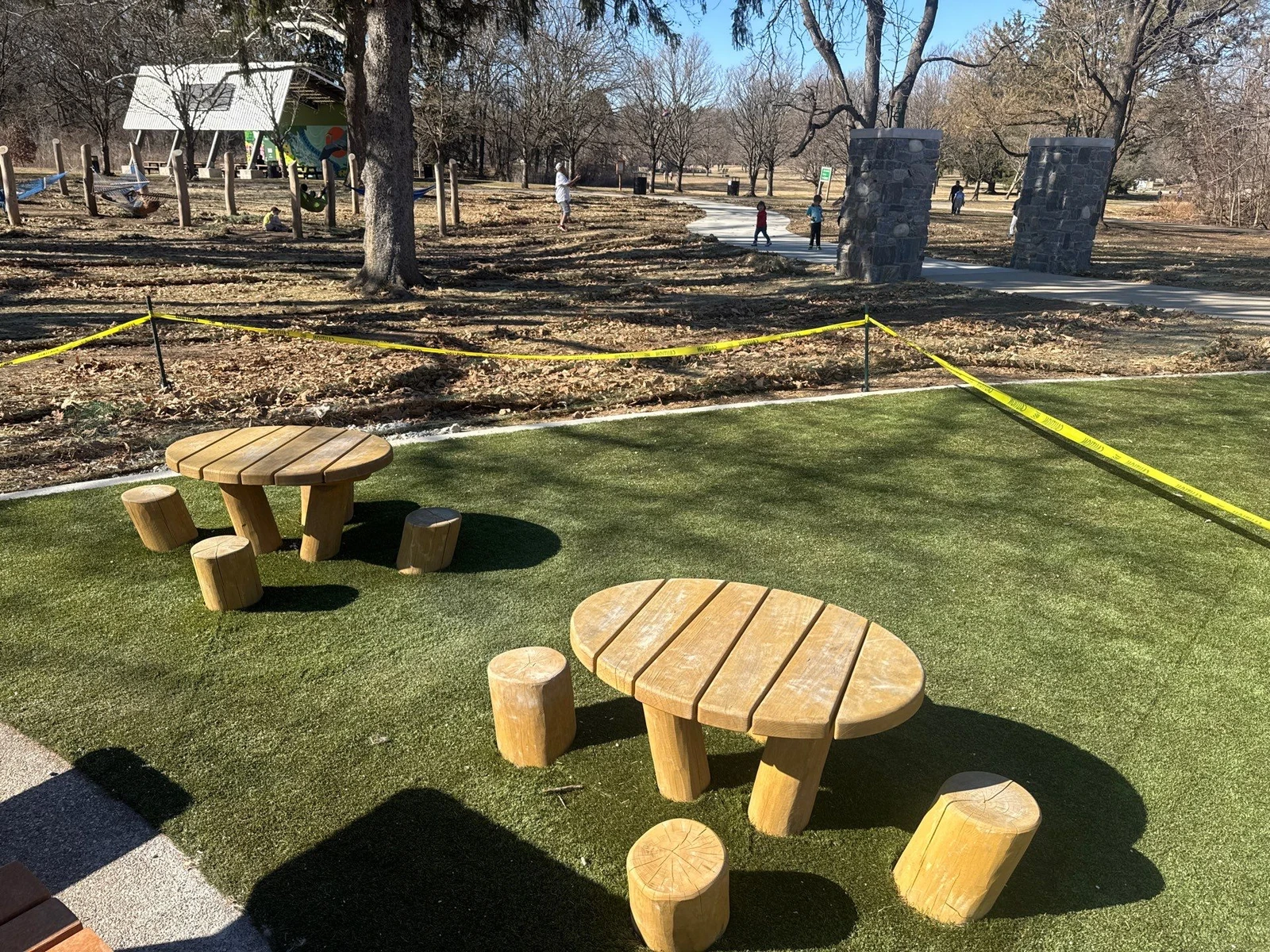 Artificial turf playground area with log-style tables and stools at Holliday Park, bordered by yellow caution tape during installation.